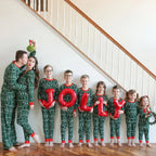 Large family of nine posing on a staircase in matching green Fair Isle holiday pajamas while kids hold red JOLLY foil balloons and parents kiss under mistletoe.