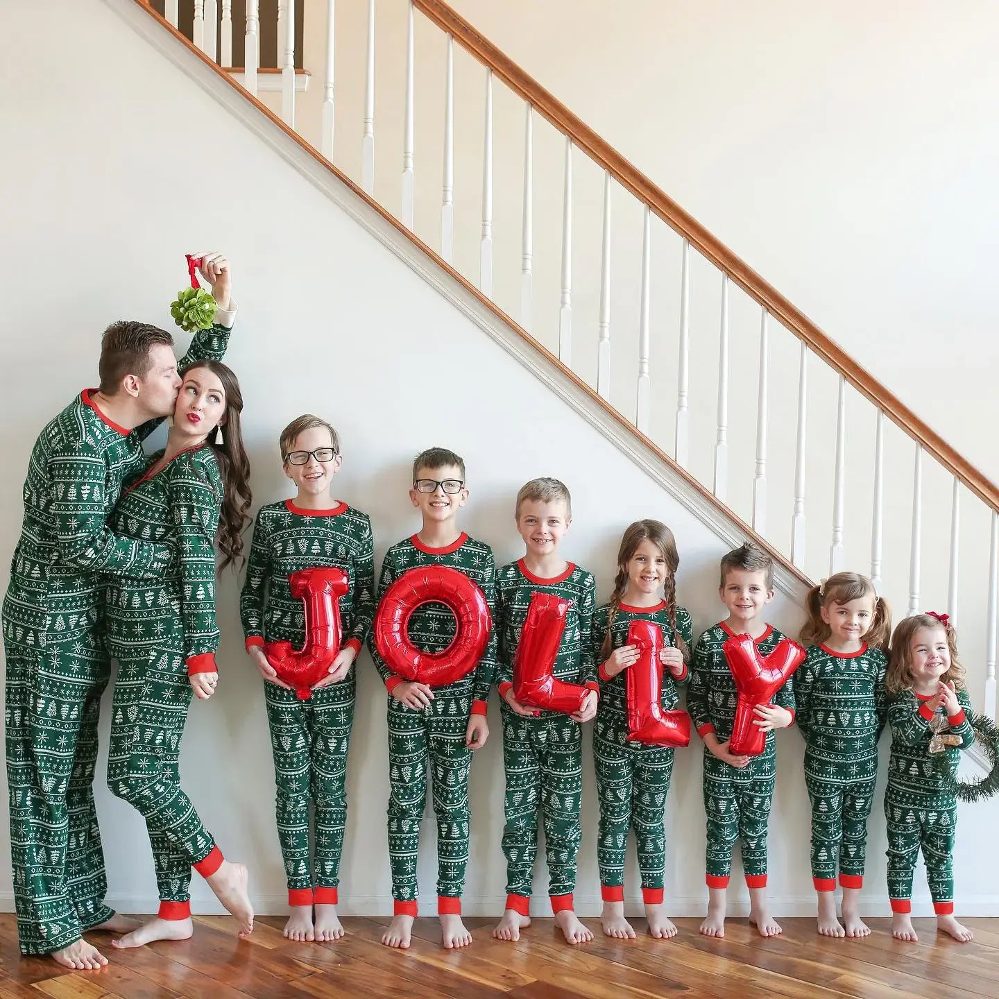 Large family of nine posing on a staircase in matching green Fair Isle holiday pajamas while kids hold red JOLLY foil balloons and parents kiss under mistletoe.