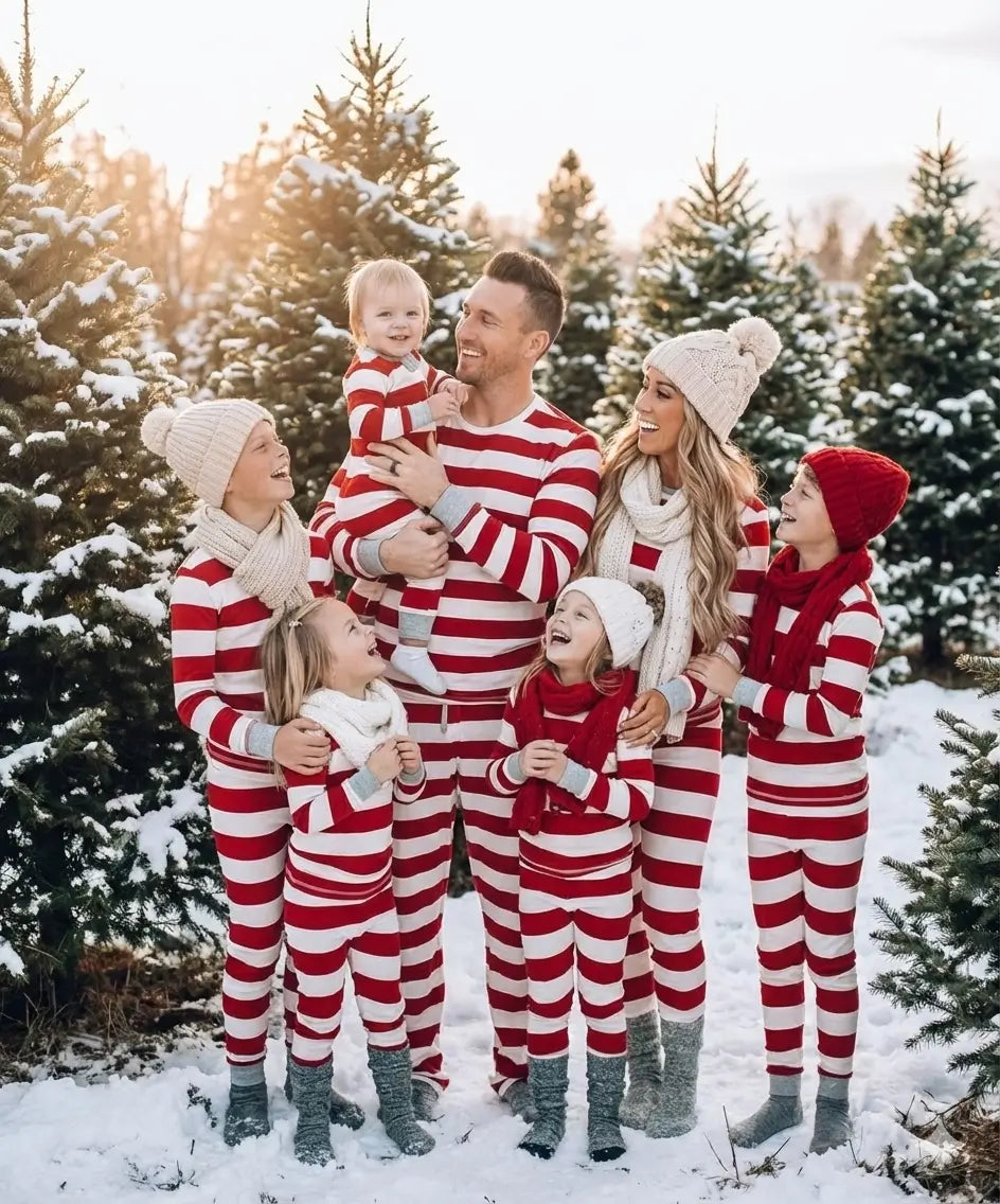 Large family of seven standing outdoors in a snowy pine tree farm wearing matching red and white striped holiday pajamas with knit beanies and scarves.