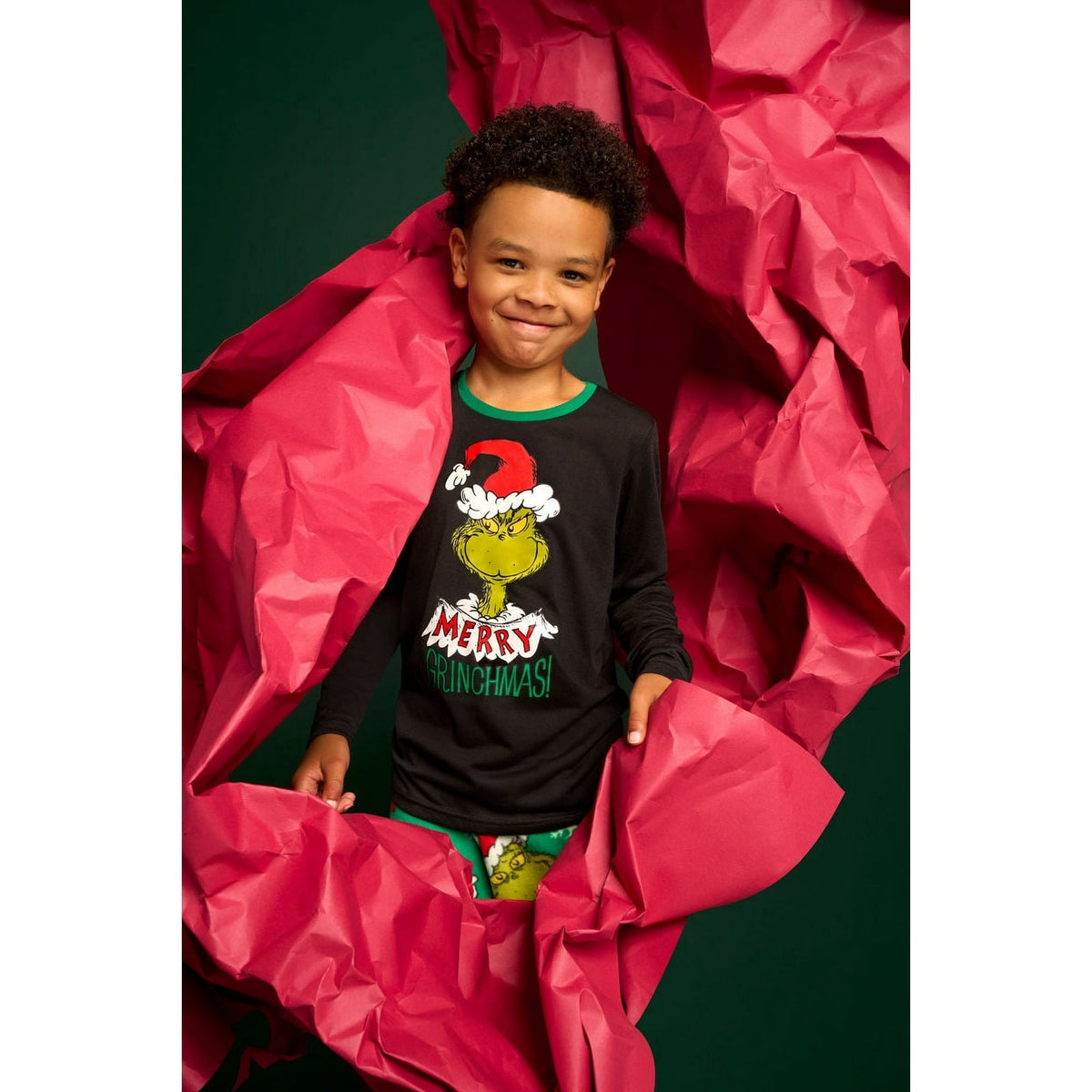 Child in a black Grinch Christmas pajama top and green printed pants surrounded by red paper