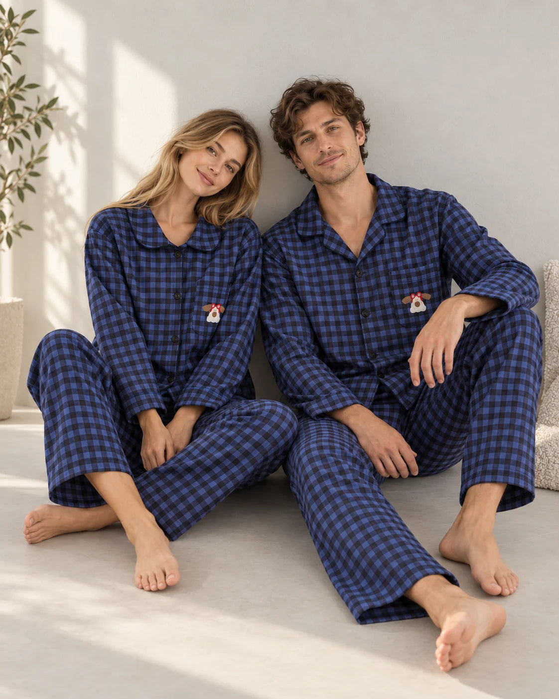 Couple seated on the floor in matching blue and black plaid pajamas, showing long sleeves, dog chest embroidery and relaxed full-length pants.