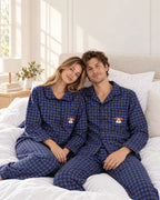 Bedroom portrait of a couple in blue plaid pajamas, with black buttons and white dog embroidery with red glasses on the chest pockets.
