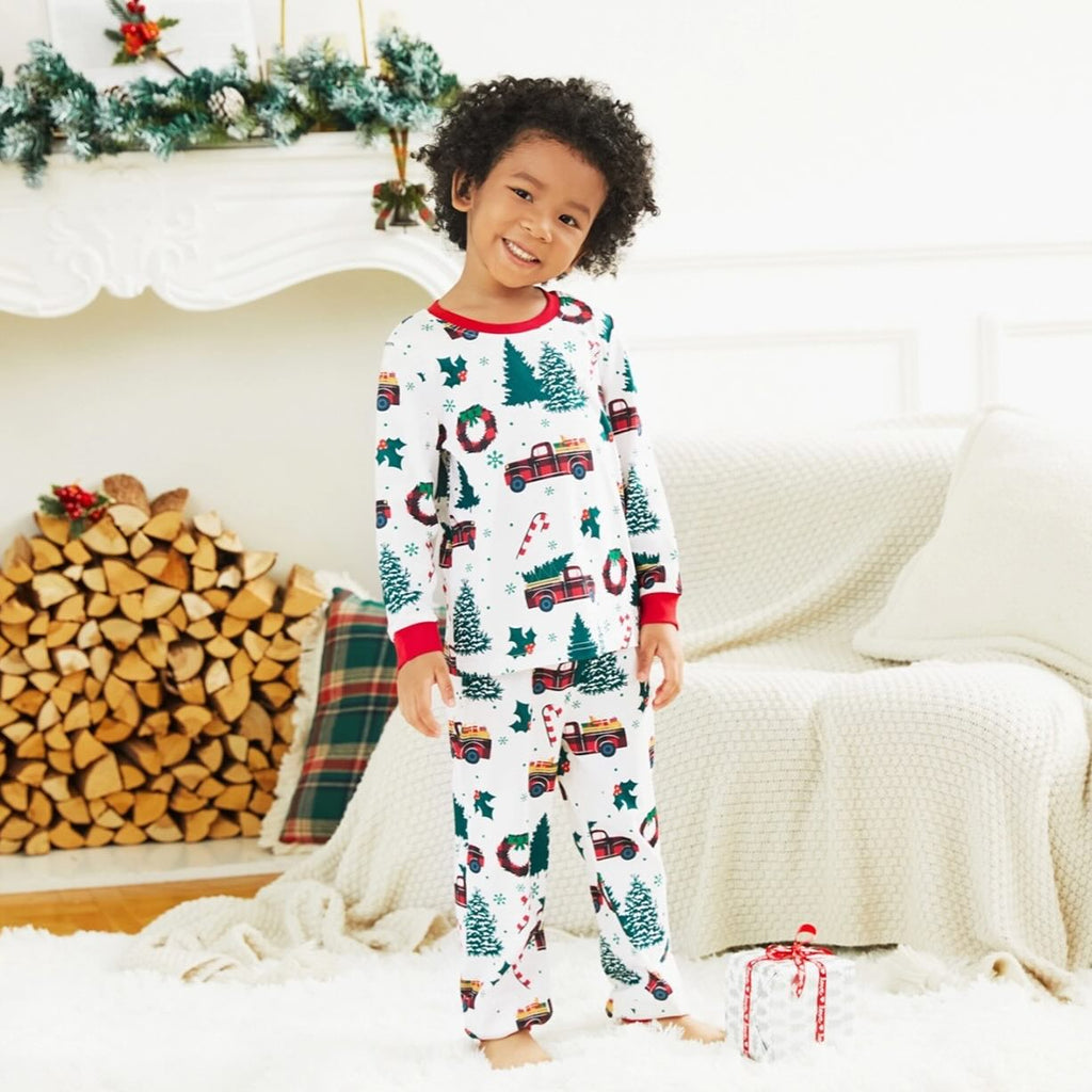 Happy young boy wearing kids matching Christmas pajamas with red truck and pine tree print in a cozy living room.