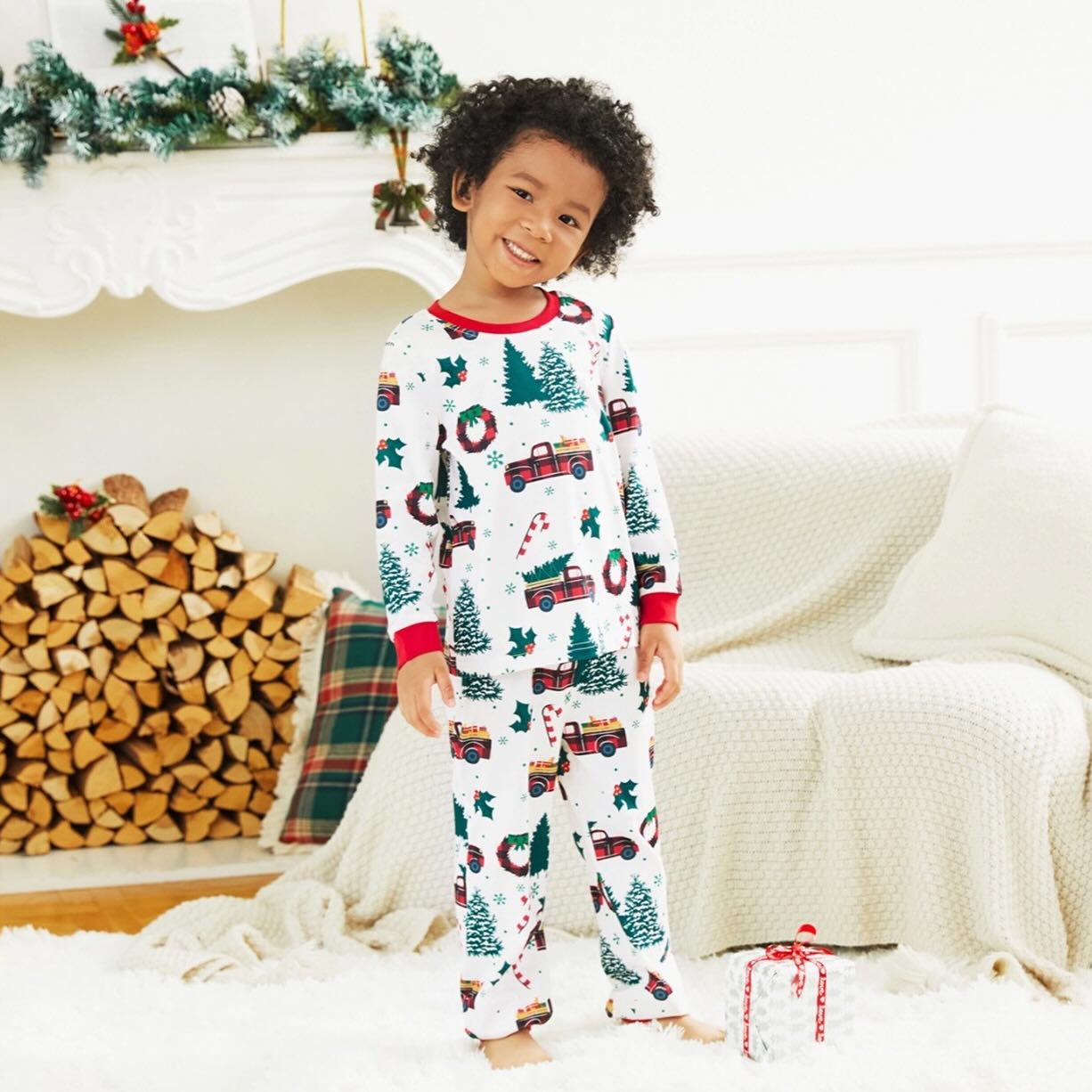 Happy young boy wearing kids matching Christmas pajamas with red truck and pine tree print in a cozy living room.