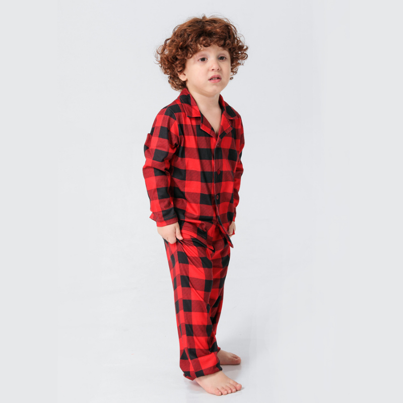 Boy with curly hair wearing red and black buffalo plaid pajamas, standing with hands in his pockets.