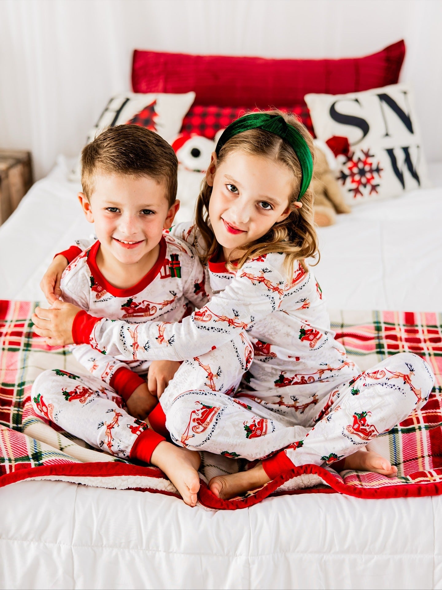 Young boy and girl with a green headband hugging on a plaid blanketed bed wearing matching Santa and reindeer pajamas.