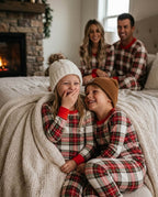 Two kids laughing together on a bed wrapped in a knit blanket wearing matching red and green plaid Christmas pajamas, parents smiling in background by fireplace.