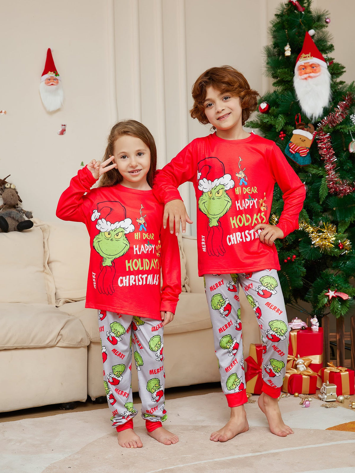 Two children wearing red Grinch tops and light gray printed Christmas pajama pants beside a tree