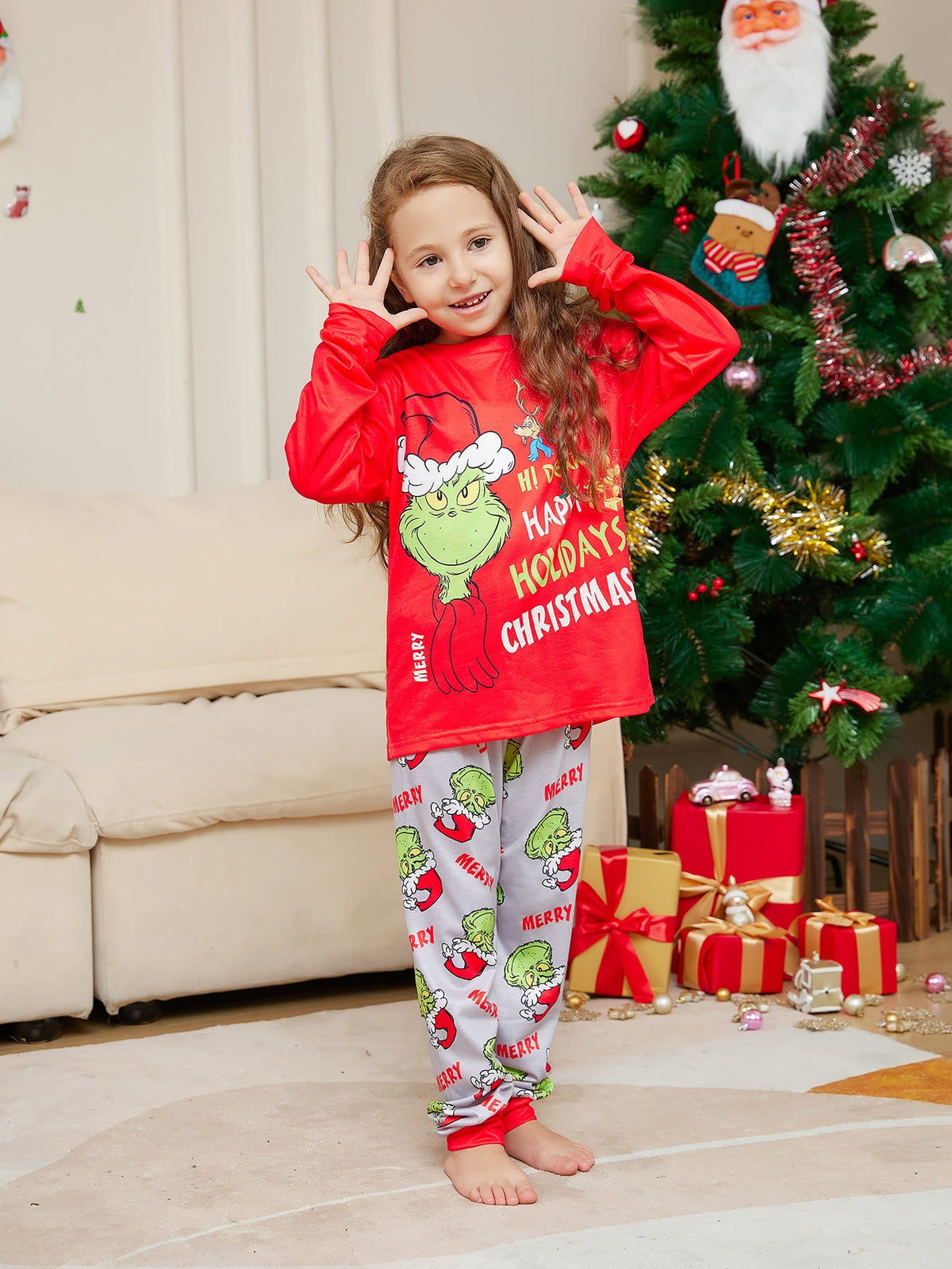Girl in a red Grinch top and light gray printed pajama pants posing beside a Christmas tree