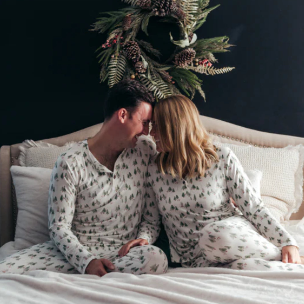 Couple sitting face to face on a bed in matching white pajamas with watercolor Christmas tree prints.