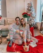 Smiling couple wearing glasses sitting on a red blanket near a flocked Christmas tree in matching white Fair Isle christmas pajamas holding coffee mugs.