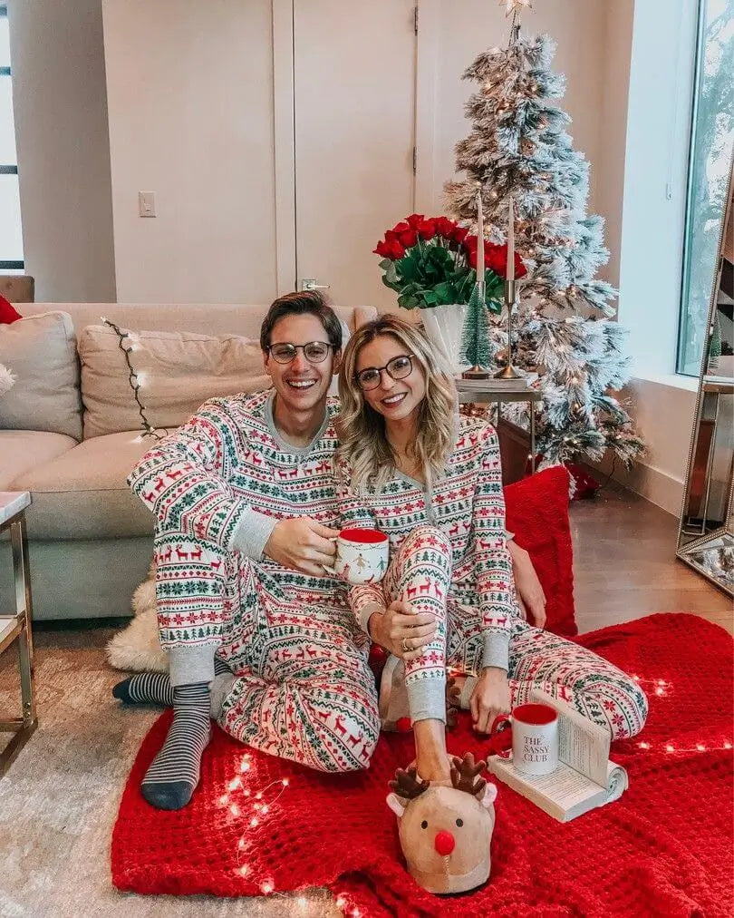 Smiling couple wearing glasses sitting on a red blanket near a flocked Christmas tree in matching white Fair Isle christmas pajamas holding coffee mugs.