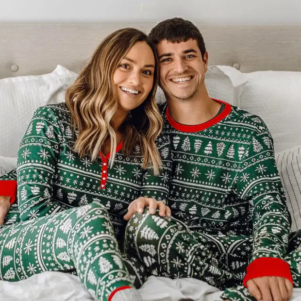 Smiling couple sitting together on a bed wearing matching green Fair Isle Christmas pajamas with red trim and henley neckline.