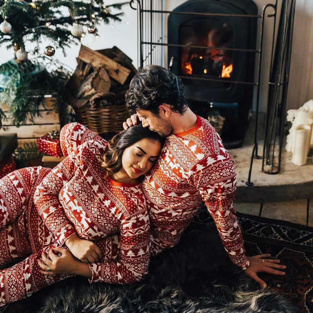 A couple cuddling on a dark faux fur rug in front of a warm lit fireplace, wearing matching red and white Nordic Fair Isle sleepwear sets.