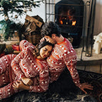 A couple cuddling on a dark faux fur rug in front of a warm lit fireplace, wearing matching red and white Nordic Fair Isle sleepwear sets.