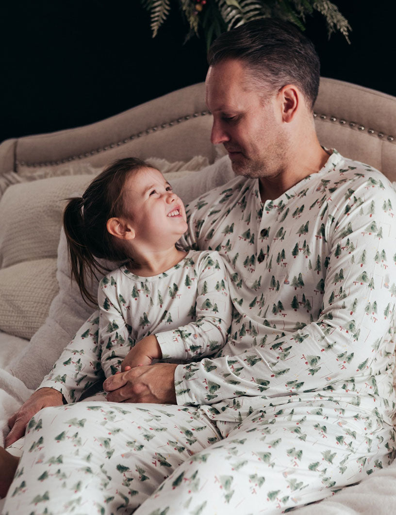 A father and his little daughter smiling at each other on a cozy bed, wearing matching white two-piece holiday loungewear sets decorated with watercolor pine trees.
