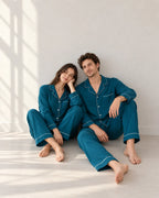 Couple seated on the floor in deep sea blue long-sleeve pajamas with white piping, chest pockets and matching full-length pants.