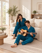 Couple relaxing in a living room, with the man playing an acoustic guitar in matching deep sea blue pajamas with white piping.