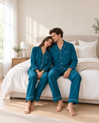 Couple sitting on a white bed in matching deep sea blue pajamas with notched collars and white piping on the cuffs and pant hems.