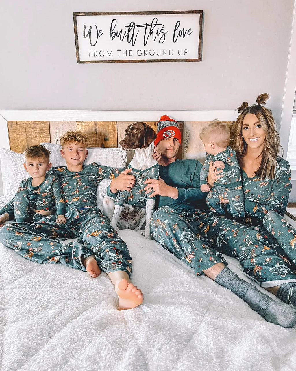 Family of five with their pet dog sitting together on a white bed wearing matching dark green safari animal printed Christmas pajamas.