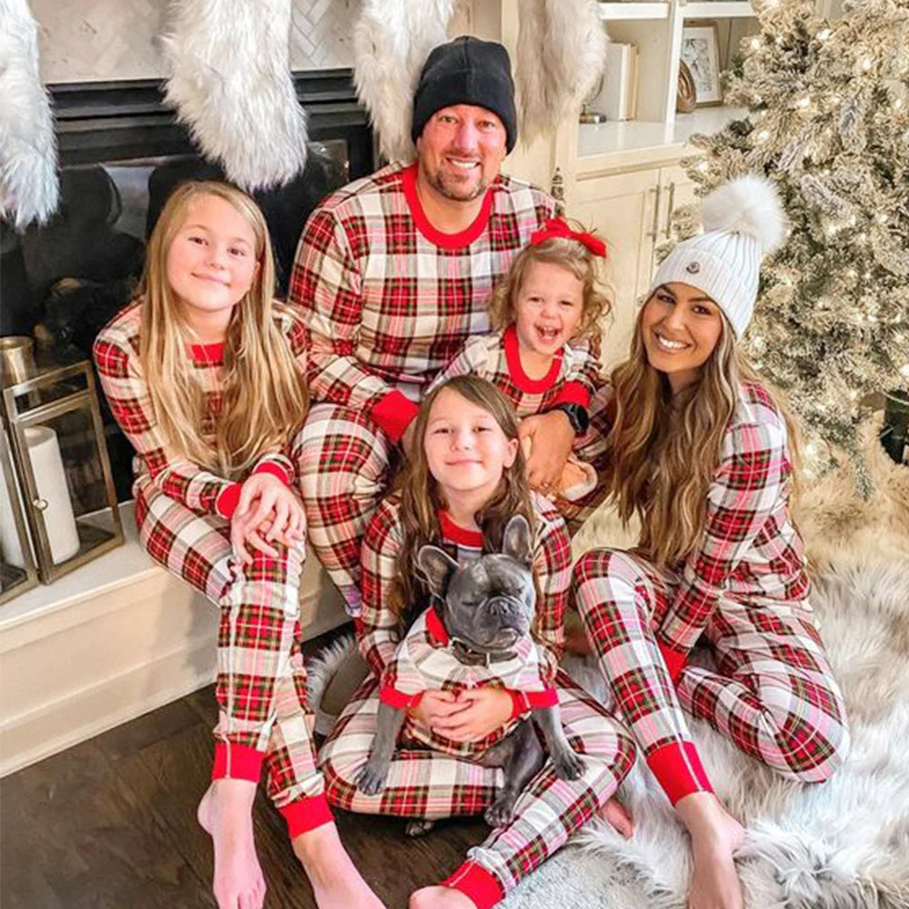 Family of five with their French bulldog wearing matching cream, red, and green plaid holiday pajamas sitting together by a fireplace and Christmas tree.