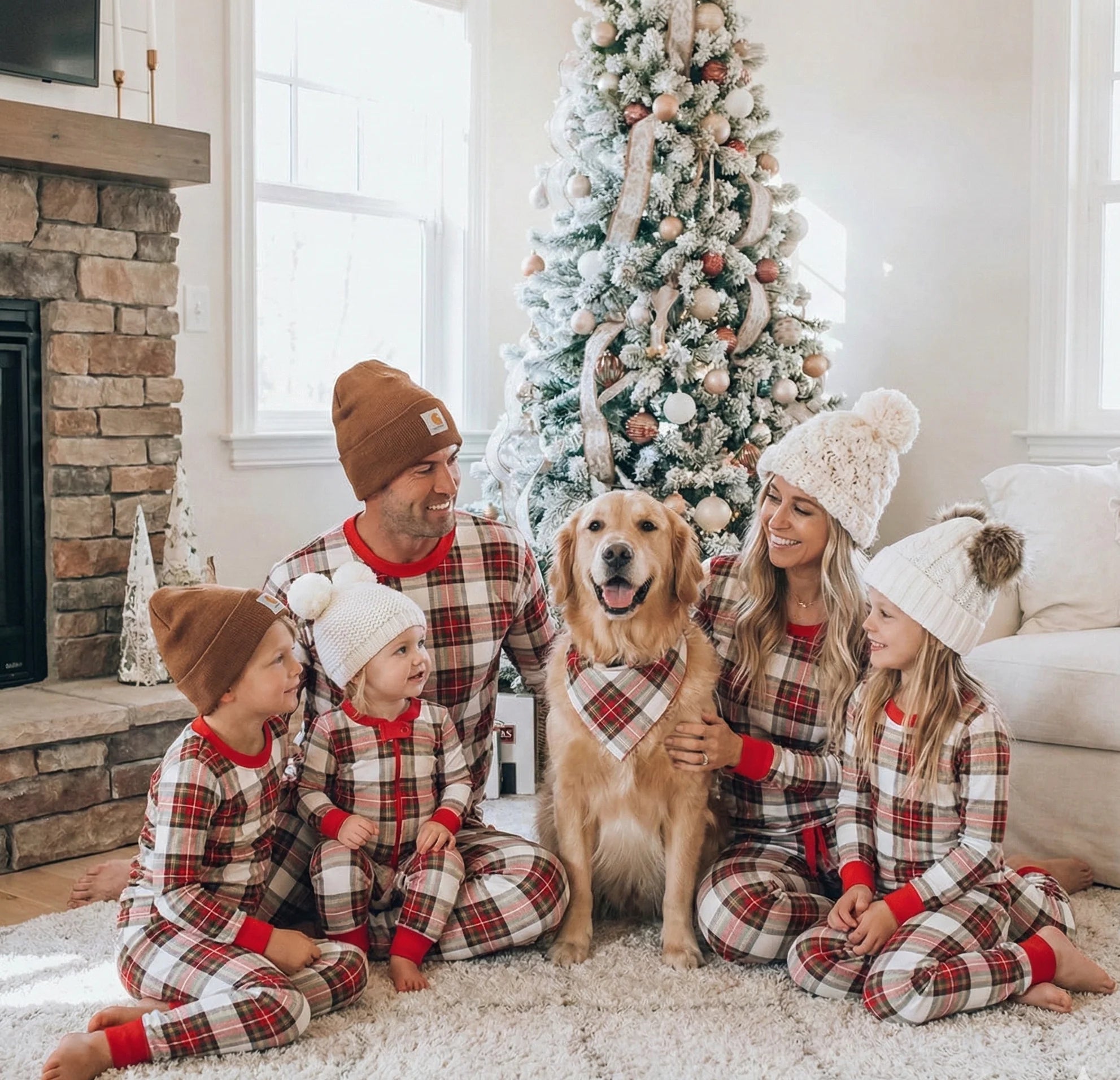 Family of five wearing winter beanies and matching cream and red plaid Christmas pajamas sitting on a rug with their golden retriever in a matching pet bandana.