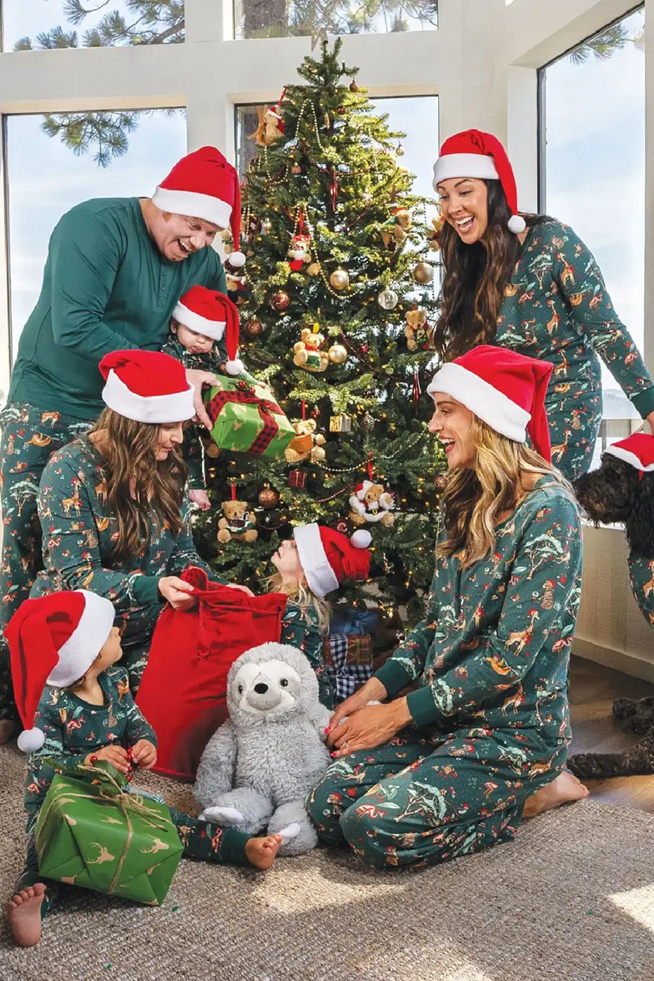 Happy family wearing red Santa hats and matching dark green safari animal pajamas opening gifts around a decorated Christmas tree.