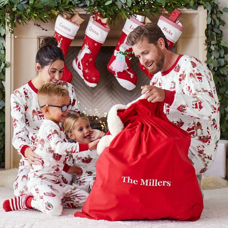 Family of four sitting on the floor gathered around a large red Santa sack near a decorated fireplace in matching Christmas pajamas.