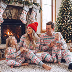 Parents, a girl and a baby sitting by a lit fireplace in grey Fair Isle Christmas pajamas beneath matching stockings.