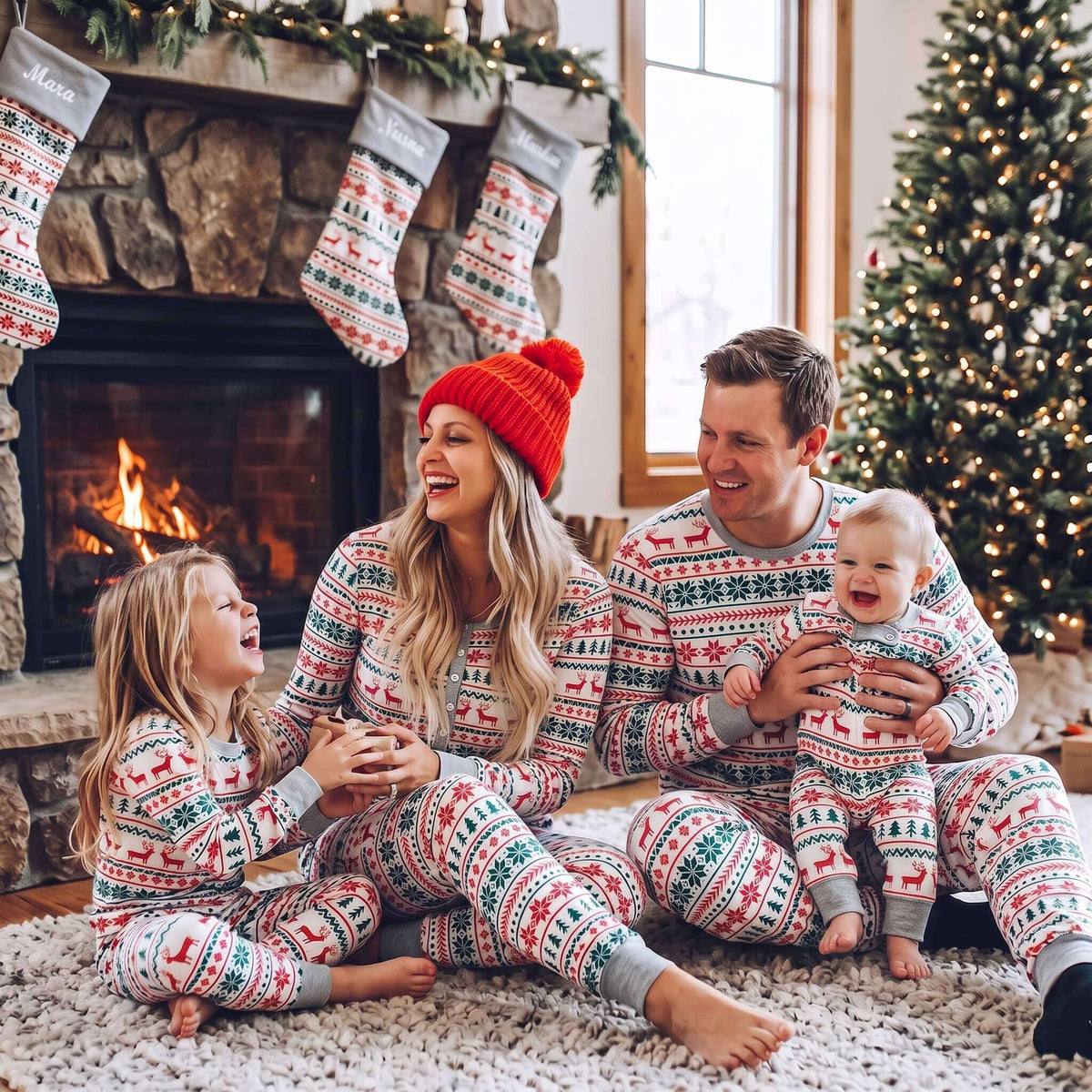 Parents, a girl and a baby sitting by a lit fireplace in grey Fair Isle Christmas pajamas beneath matching stockings.
