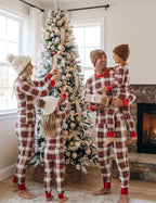 Family of four decorating a flocked Christmas tree indoors wearing matching cream and red plaid Christmas pajamas with red cuffs, fireplace in background.