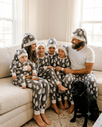 Family in grey Christmas-tree pajamas and matching hats on a sofa with a black dog, dad in a white T-shirt.