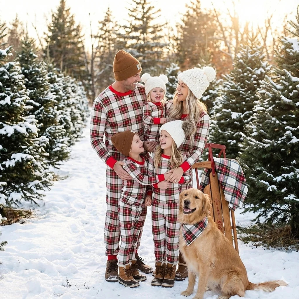 Family with three kids and a Golden Retriever wearing matching red and cream plaid holiday pajamas at an outdoor snow-covered Christmas tree farm with a wooden sled.