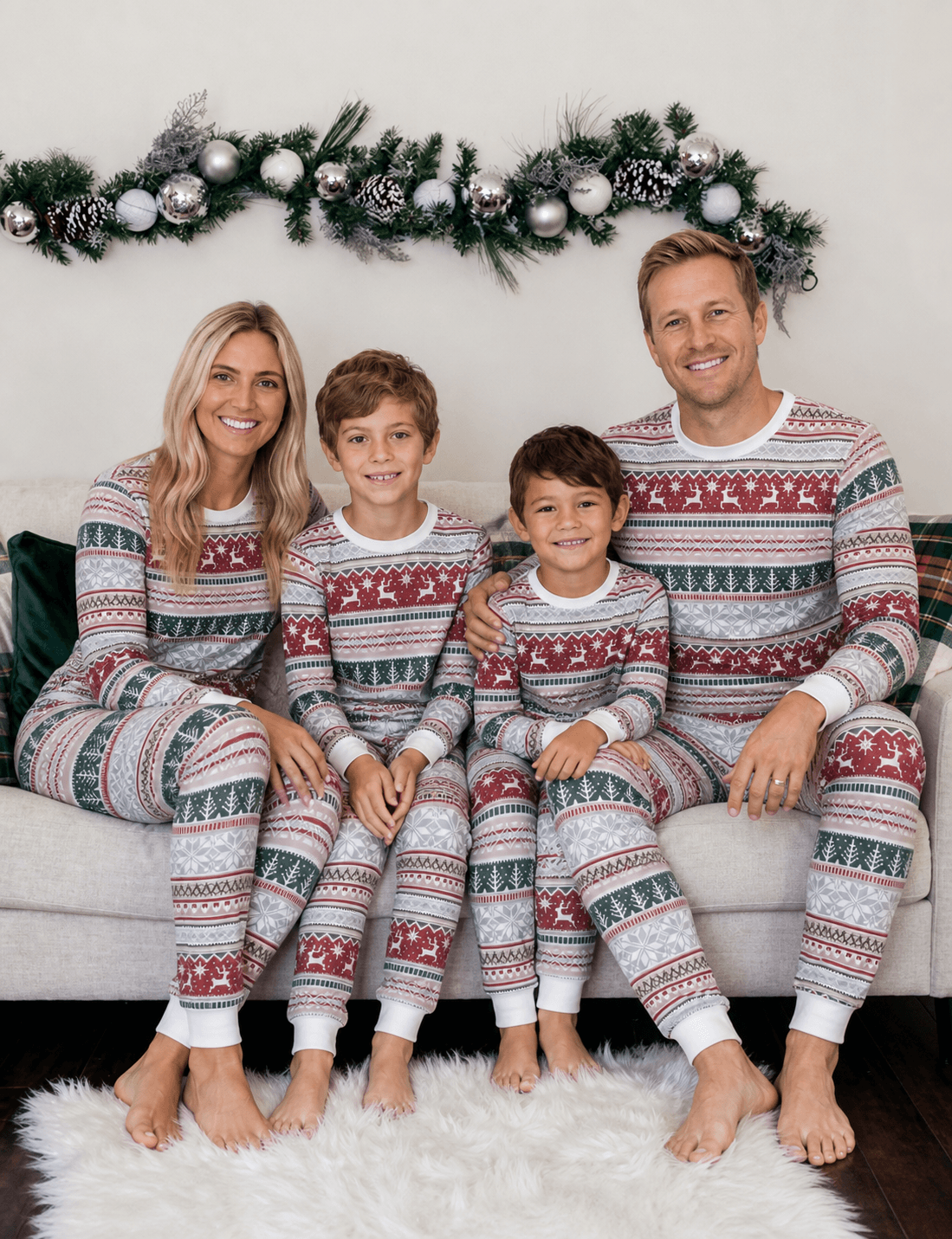 Family sitting on a sofa beneath a holiday garland in grey Christmas pajamas with red reindeer and green trees.