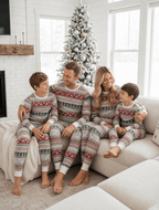Family on a cream corner sofa in light grey Christmas pajamas with red reindeer and green trees, beside a snowy tree.