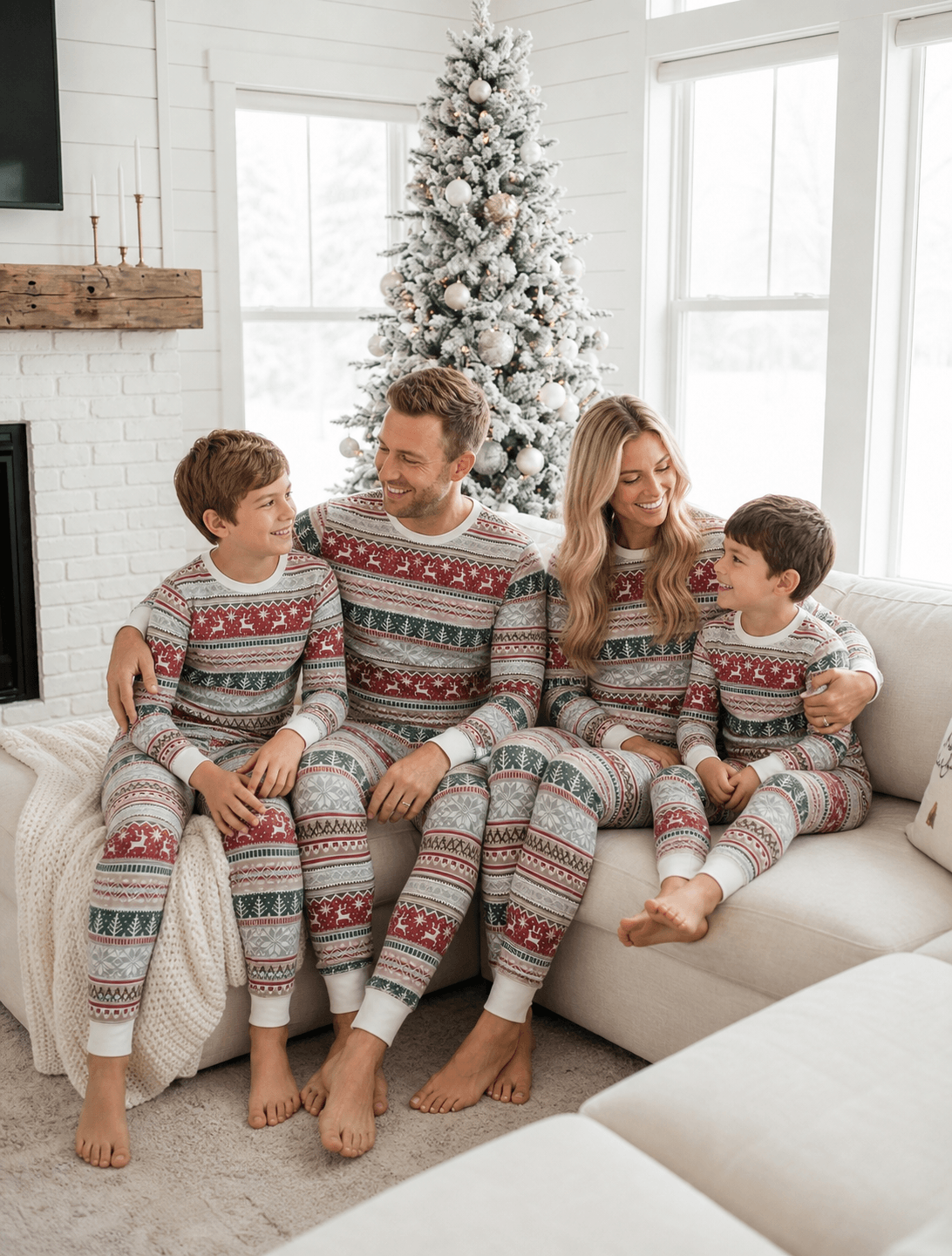 Family on a cream corner sofa in light grey Christmas pajamas with red reindeer and green trees, beside a snowy tree.