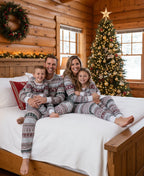 Smiling family of four sitting on a white bed inside a cozy log cabin wearing matching grey Fair Isle Christmas pajamas with burgundy reindeer patterns.