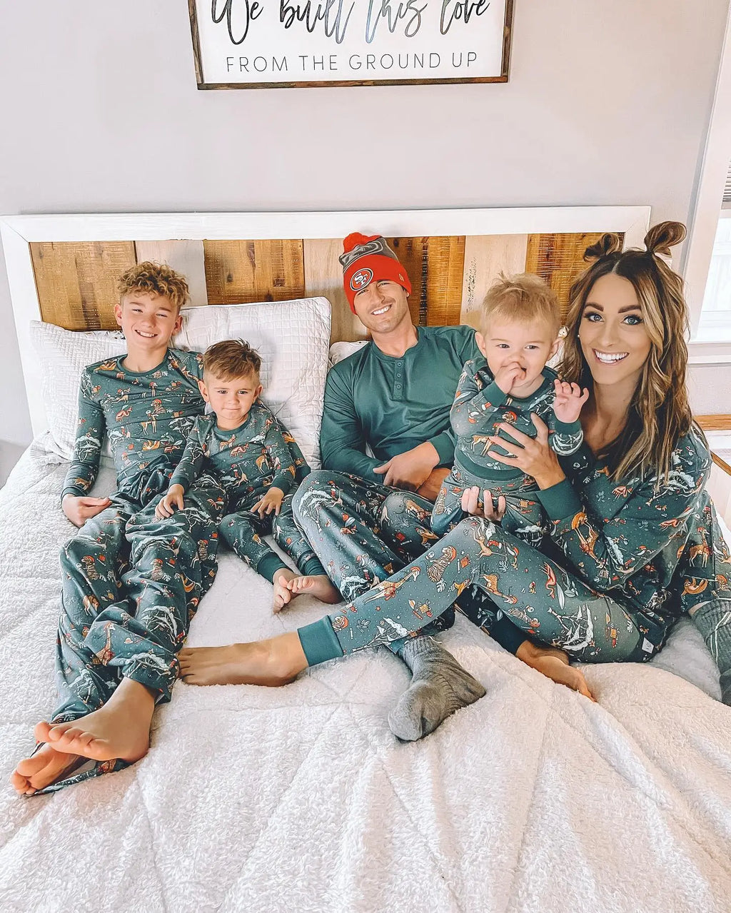 Happy family of five wearing matching dark green safari Christmas pajamas relaxing together on a white bed.