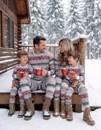 Family in matching grey Fair Isle pajamas with winter boots sitting on snow-covered cabin porch holding mugs.