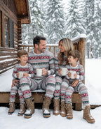 Family in matching grey Fair Isle pajamas with winter boots sitting on snow-covered cabin porch holding mugs.