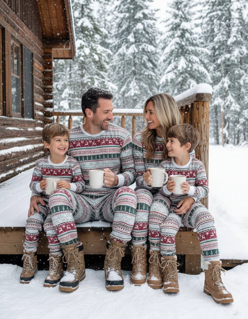 Family in matching grey Fair Isle pajamas with winter boots sitting on snow-covered cabin porch holding mugs.