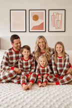 Family sitting together on a cream bed in matching red, white and black plaid Christmas pajamas with red trim.