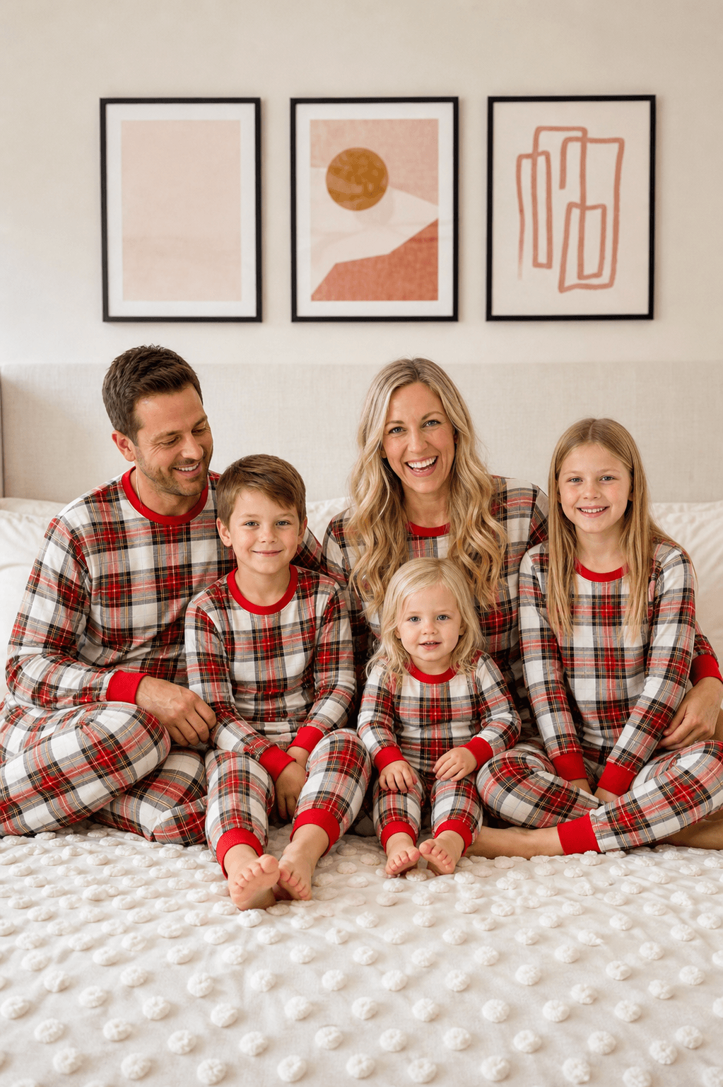 Family sitting together on a cream bed in matching red, white and black plaid Christmas pajamas with red trim.