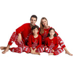 Family of four sitting cross-legged in a row on a white floor, wearing matching red Christmas pajamas with white brush-stroke tree graphics and Nordic patterned sleepwear bottoms.