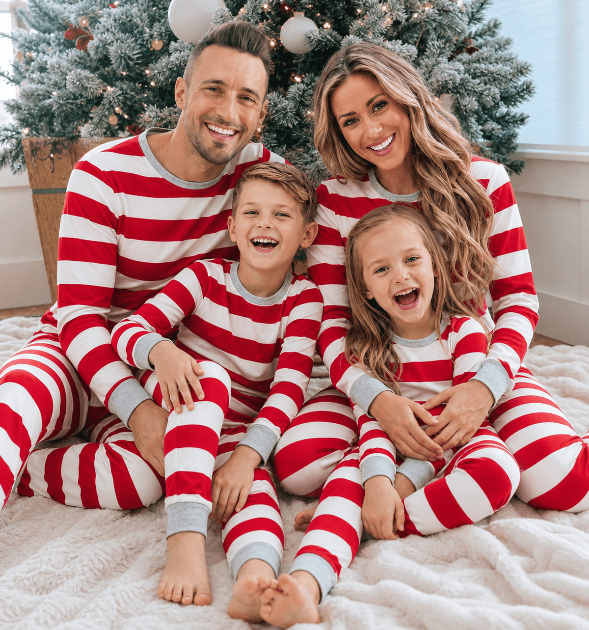 Parents and two children laughing by a Christmas tree in pajamas with wide red and white stripes and grey trim.