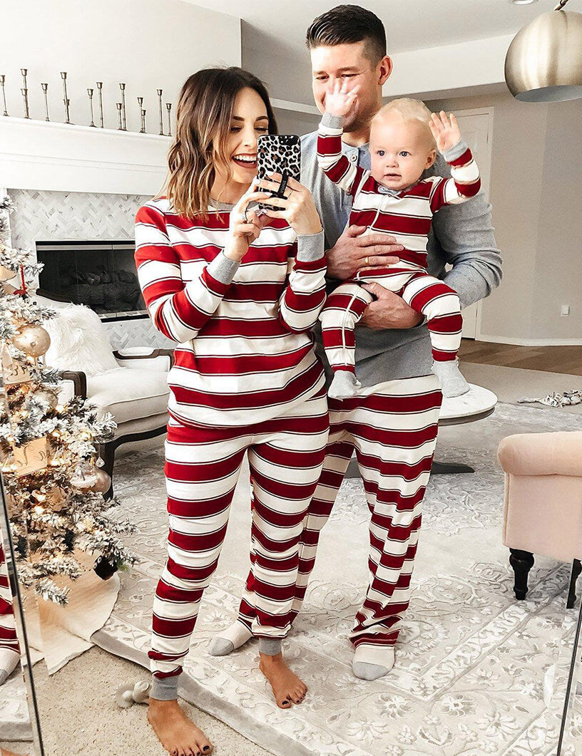 Parents and baby taking a mirror selfie wearing matching red and white candy cane striped Christmas pajamas with grey cuffs near a festive tree.
