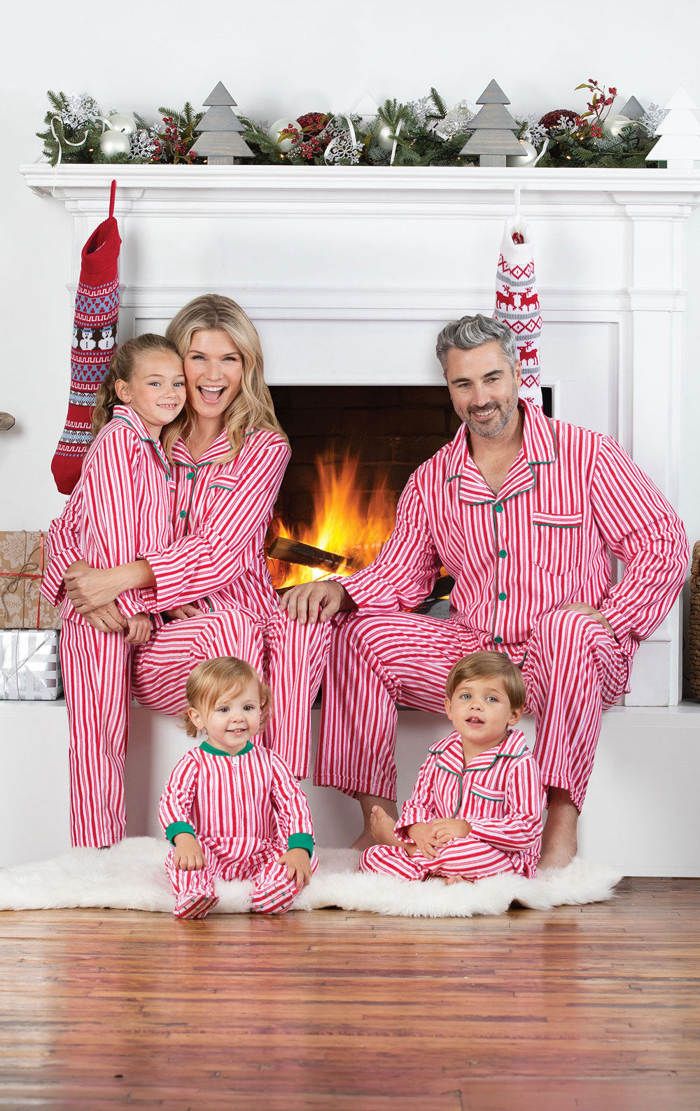 A family of five sitting in front of a warm lit fireplace wearing matching red and white candy cane striped holiday pajamas.