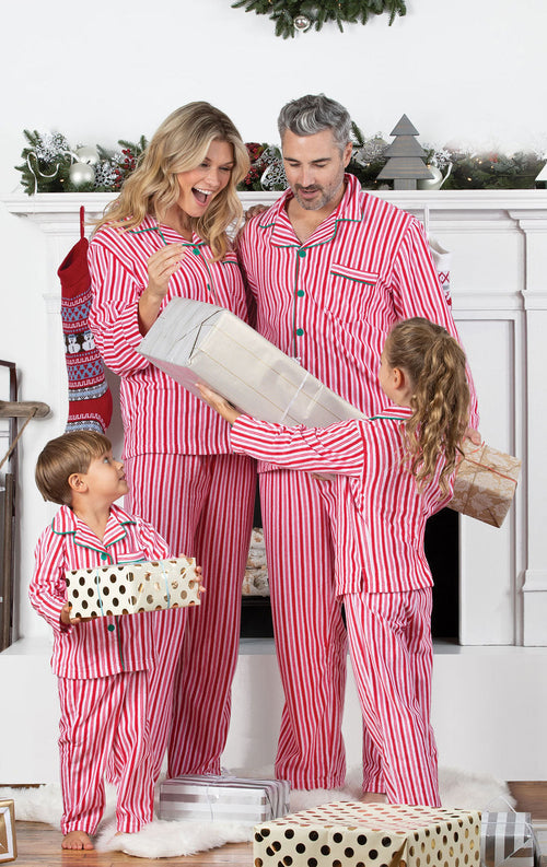 Family of four standing together in front of a glowing Christmas tree with present boxes, wearing matching red and white candy cane striped pajamas.
