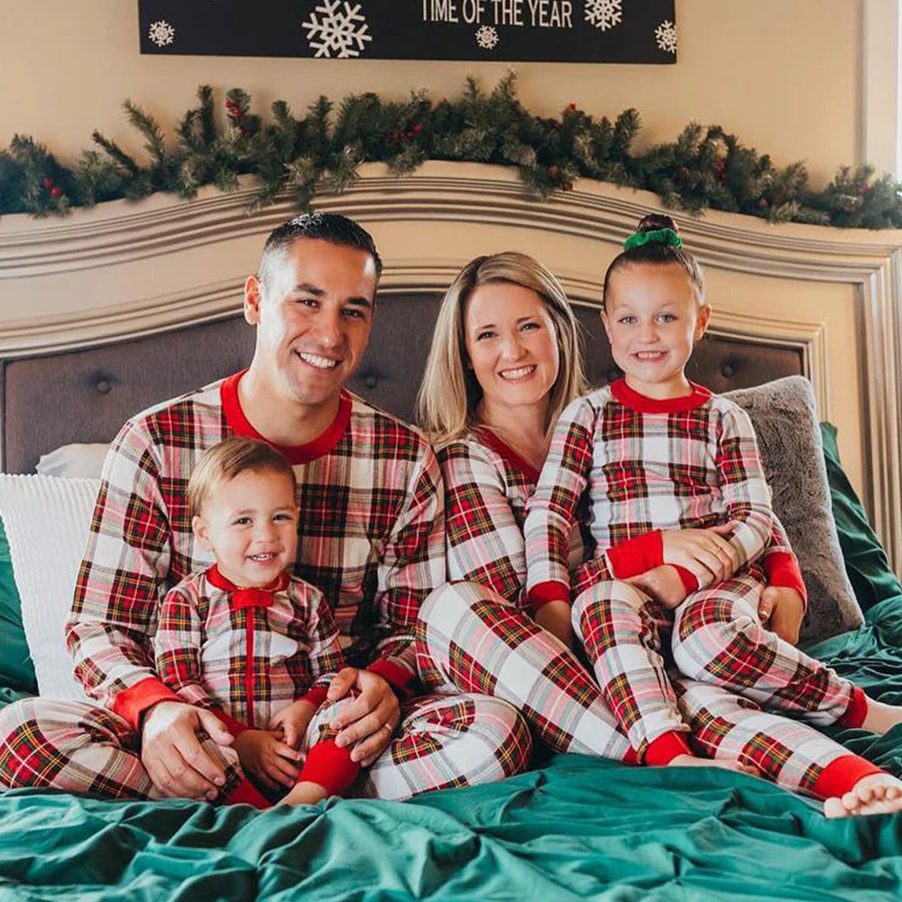 Family of four sitting on a green bed in matching cream and red plaid pajamas, featuring parents with a toddler and young daughter smiling.