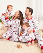 Mother, father, toddler, and baby sitting on a plush rug in matching white Santa and reindeer print pajamas holding small holiday gifts.